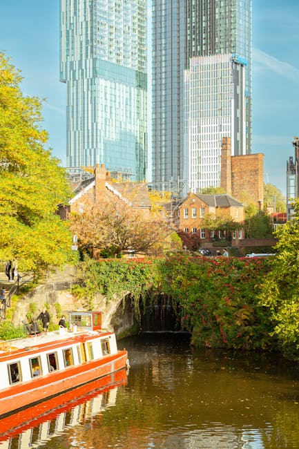 A city scene featuring a river with a red and white boat cruising along calm waters, surrounded by lush green trees with some foliage displaying autumn colors. In the background, there are traditional